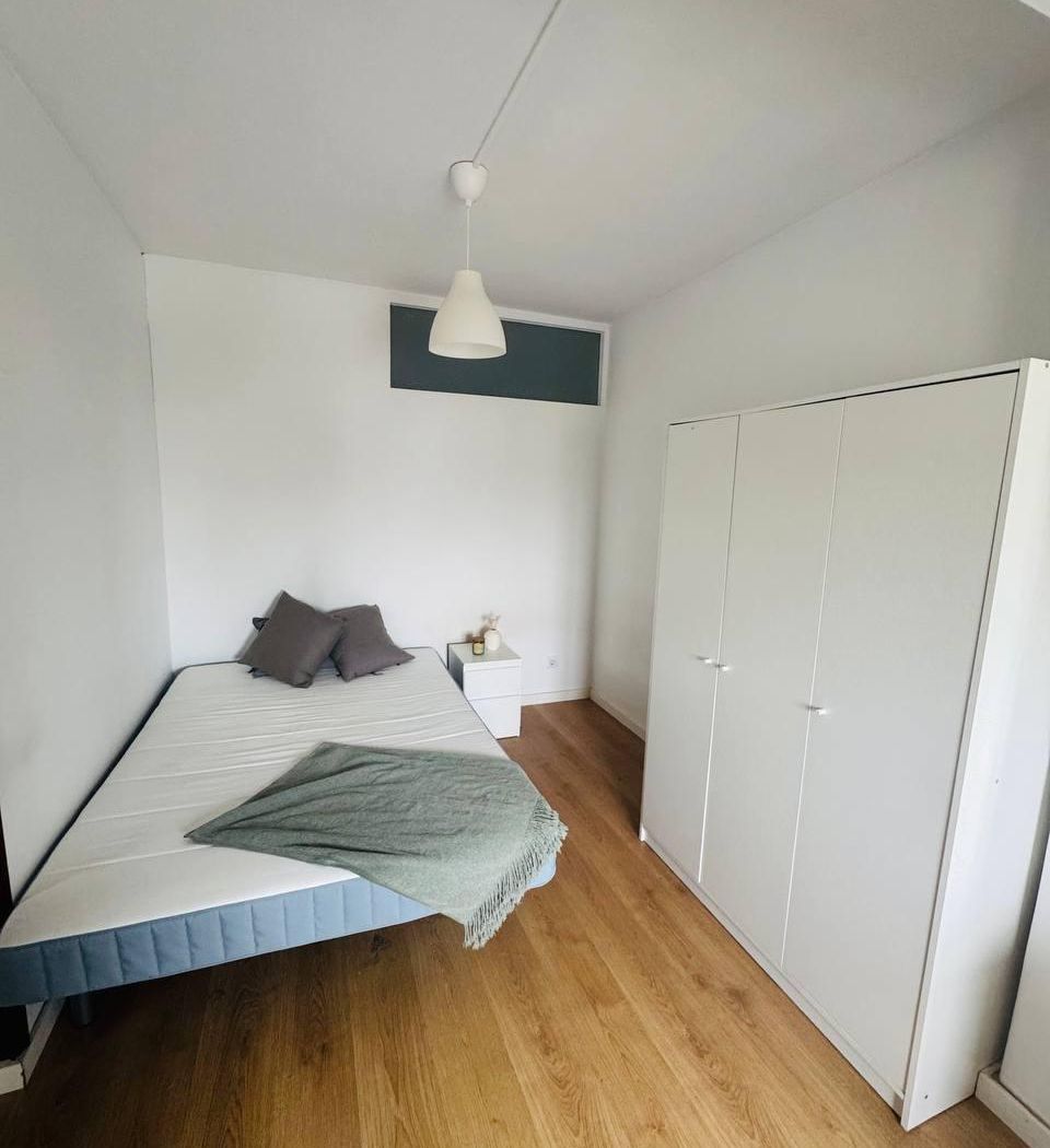 The room seen from the door: wardrobe, full-length mirror, chest of drawers, desk, and a window over the rooftops.